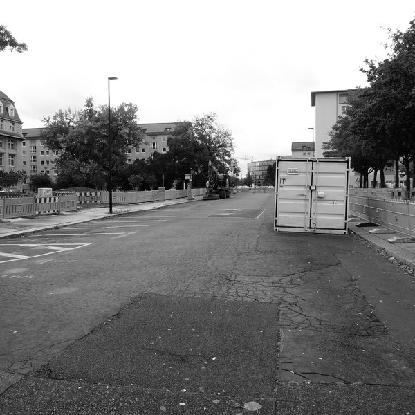 Abgesperrter Straßenbereich, in dem ein Bagger parkt und ein weißer Werkzeug-Container abgestellt sind. Quadratische Schwarz-Weiß-Aufnahme.
