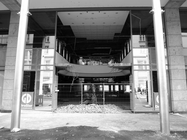 Schwarz-Weiße Panorama-Aufnahme der Glasfassade, aus der der Bereich des Eingangs und ein Stockwerk darüber, herausgerissen wurde, inkl. eines Teils der Böden.