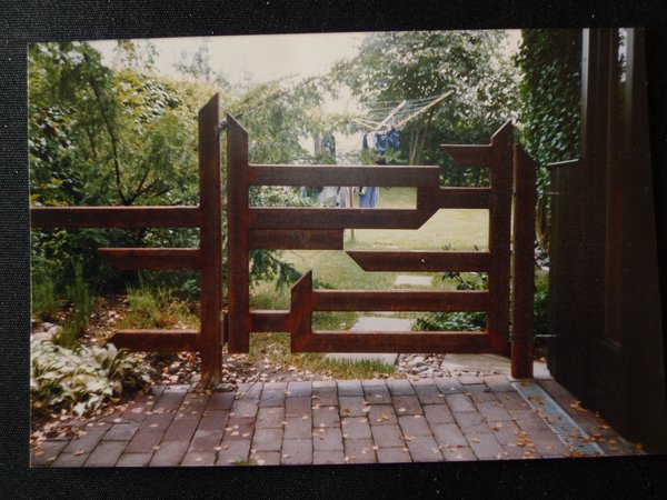 Ein "Freiform"-Tor aus massiven Träger, die in sehr grafischer Form zueinander angeordnet sind, steht am Eingang zu einem Garten, in dessen Hintergrund eine Wäschespinne auf der Wiese zu sehen ist.