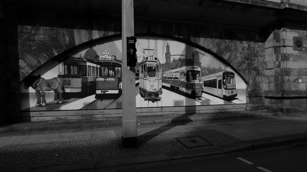 Von links nach rechts sind veschiedene Straßenbahn-Generationen abgebildet: links mit einem Pferd als "Pferdestärke", rechts daneben vier weitere Generationen. Im Hintergrund sind verschiedene historische Gebäude angedeutet: Yenidze, Hofkirche, Hausmannsturm.
