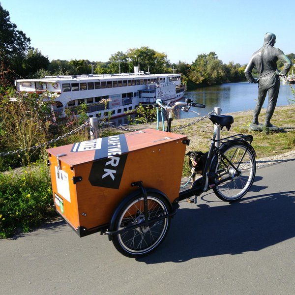 Auf dem Elbradweg steht das Lastenrad, mit großer Transportbox vor dem Lenker. Links im Hintergrund: das Hotelschiff an seinem Anlegeplatz, rechts eine Seefahrer-Statue (die inzwischen nicht mehr dort aufgebaut ist). Quadratische Farbaufnahme.