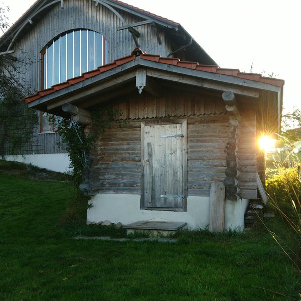 Sicht auf die kleine Hütte im Vordergrund, und das Haupthaus links im Hintergrund. Rechts an der Außenwand scheint die frühe Morgensonne hervor.