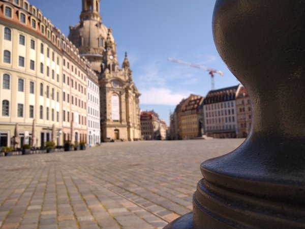 Beobachter-Perspektive, seitlich hinter einem Poller, der zum Eingang des Neumarktes führt, scharf rechts im Vordergrund, während der Neumarkt in der Tiefenunschärfe verschwimmt.
