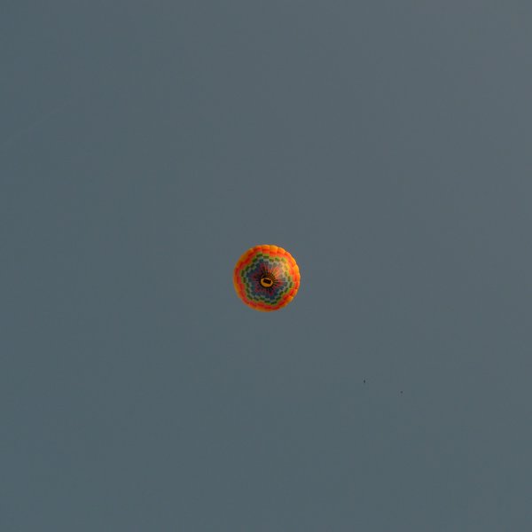 Inmitten einer quadratischen Farbaufnahme ist klein ein Heißluftballon zu sehen, der direkt von unten (90°) aufgenommen ist. Der Ballon ist orange und hat im unteren Bereich regenbogen-farbene Farbfelder. Der Korb befindet sich exakt in der Mitte der Aufnahme. Der wolkenlose Morgenhimmel ist graublau.