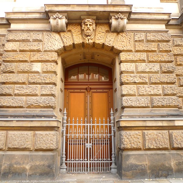Eine imposante hölzerne Außentüre ist durch ein personenhohes Gitter versperrt. Über der Tür ragt der Kopf einer Skulptur, der auf die Straße schaut.