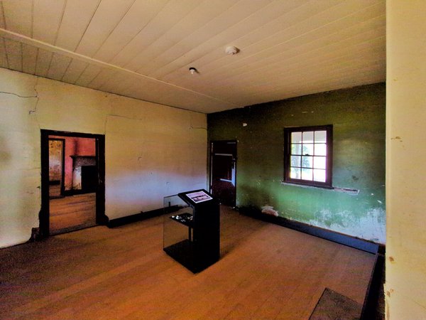 The main room of the house. It's in relatively good repair, with new-ish floorboards. The far wall is green and has a window and a heavy wooden door. The other walls and ceiling are cream-coloured.