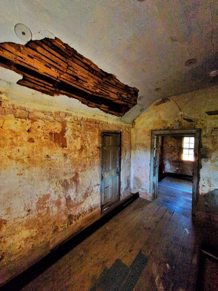 A room where the white ceiling has partially fallen down, revealing the timber beams underneath. The render has mostly come off the side wall.