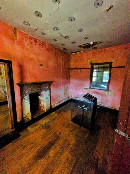 A room with faded pink walls that might have been a kitchen. The floors look like they've been restored at some point. The white ceiling has a number of transparent circular disks with bolts that are keeping it from falling down. There's a fireplace and a door on the wall on the left, and the far wall has a window into another room.