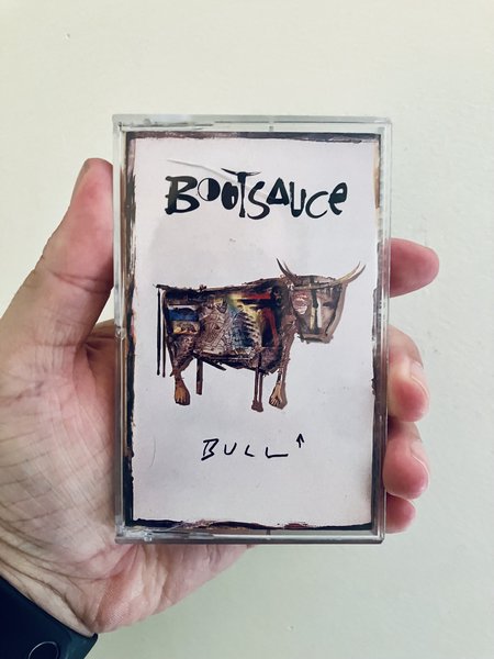 A hand holds a cassette copy of Bootsauce’s Bull against a plain light wall. The cover has a mostly white background with the stylized “Bootsauce” logo at the top, an abstract horned bull illustration in the centre, and the handwritten title “Bull” near the bottom.