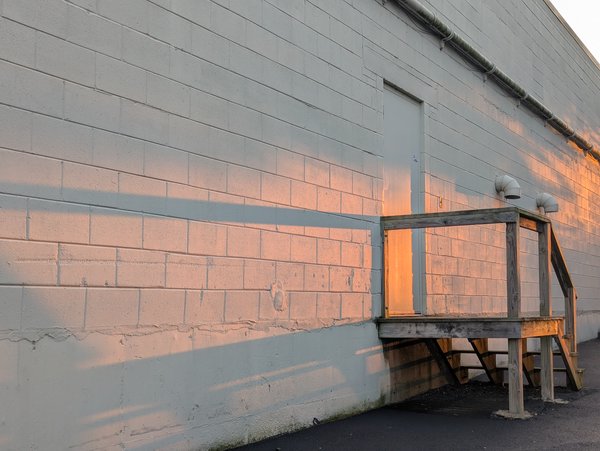 Side of an industrial building painted white with a wooden staircase with a setting sun casting warm light and long shadows.